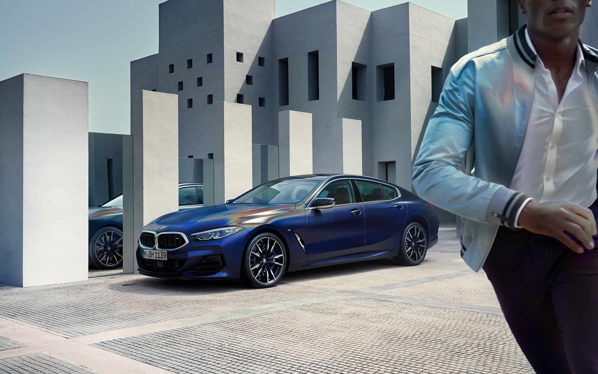 Elegant coupé silhouette with M light alloy wheels 895 M Bicolour. BMW M850i xDrive Gran Coupé G16 LCI Facelift 2022 BMW Individual Frozen Tanzanite Blue metallic three-quarter front view standing with male model in foreground