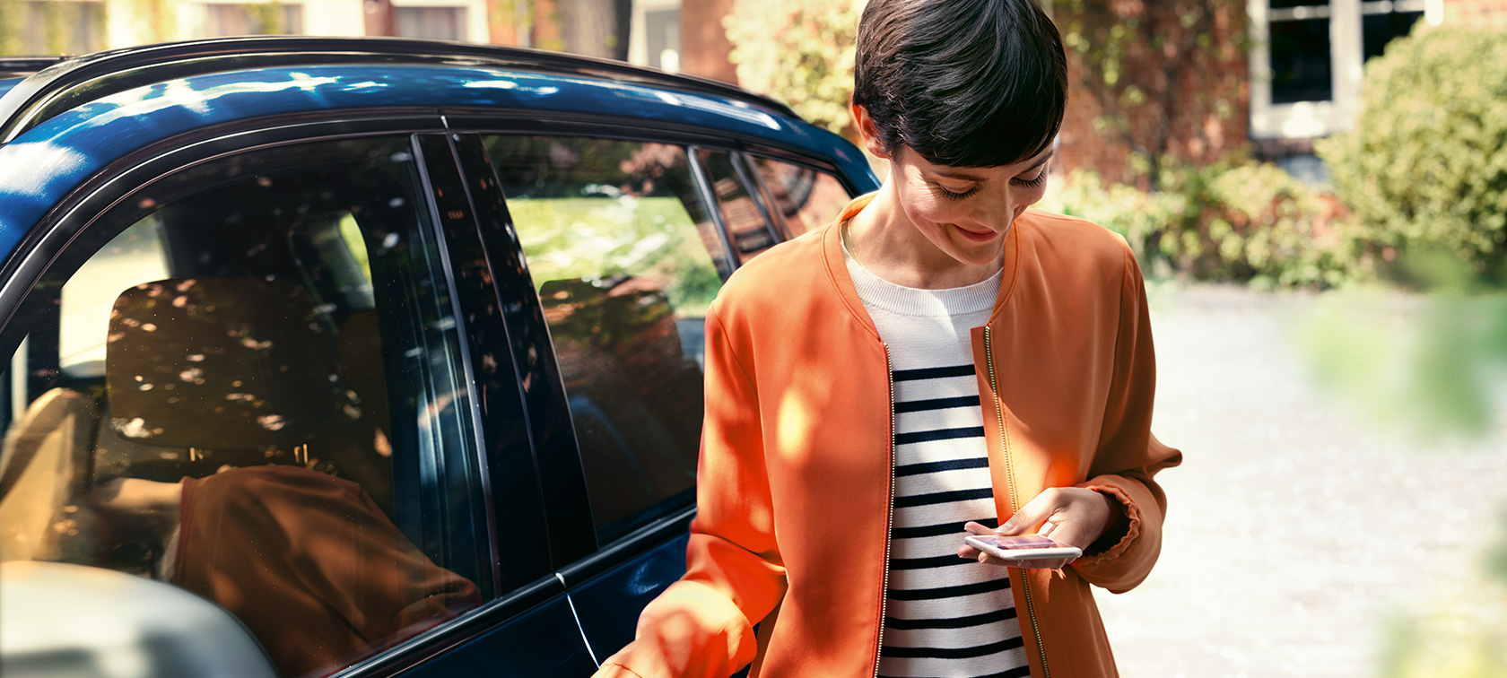 Dark-haired woman in front of BMW Dark-haired woman in orange jacket in front of BMW looking at her smartphone
