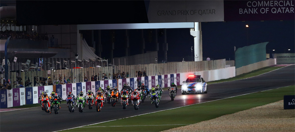 Start of the MotoGP Grand Prix of Qatar at night, with riders accelerating ahead of the BMW M Safety Car under illuminated signage