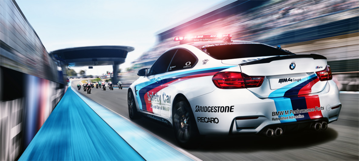BMW M4 Coupé Safety Car leading MotoGP riders out of the pit lane at high speed, featuring M Performance Parts livery and race-day atmosphere